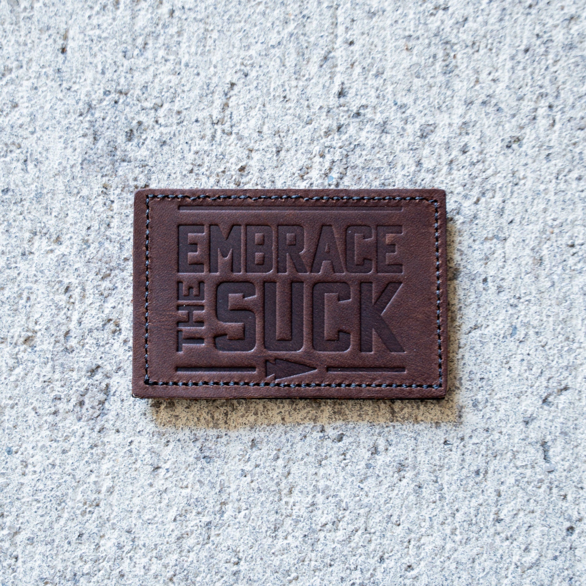The Patch - Embrace The Suck features embossed lettering on brown leather, shown on concrete—an eye-catching addition to any Velcro patch collection.
