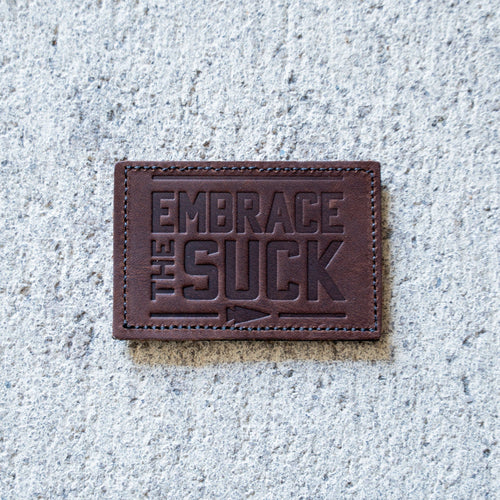 The Patch - Embrace The Suck features embossed lettering on brown leather, shown on concrete—an eye-catching addition to any Velcro patch collection.
