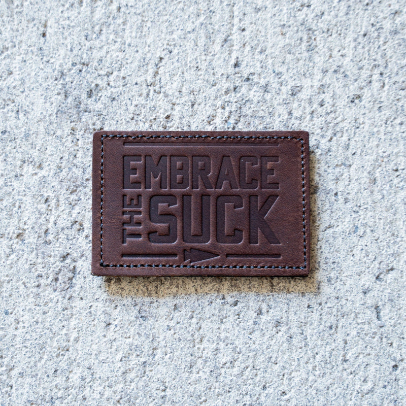 The Patch - Embrace The Suck features embossed lettering on brown leather, shown on concrete—an eye-catching addition to any Velcro patch collection.
